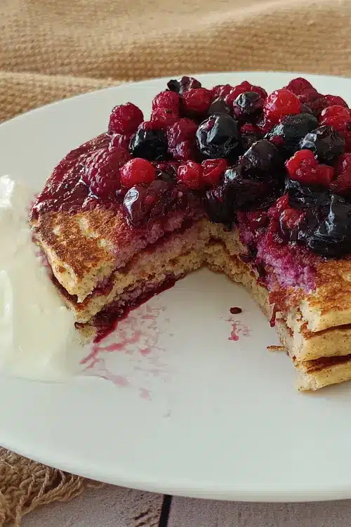 Pancakes protéinés IG bas : la recette au skyr Part entamée d'une pile de pancakes à l'avoine et au skyr, avec l'intérieur moelleux, les fruits rouges et le skyr. Pancakes protéinés IG bas.